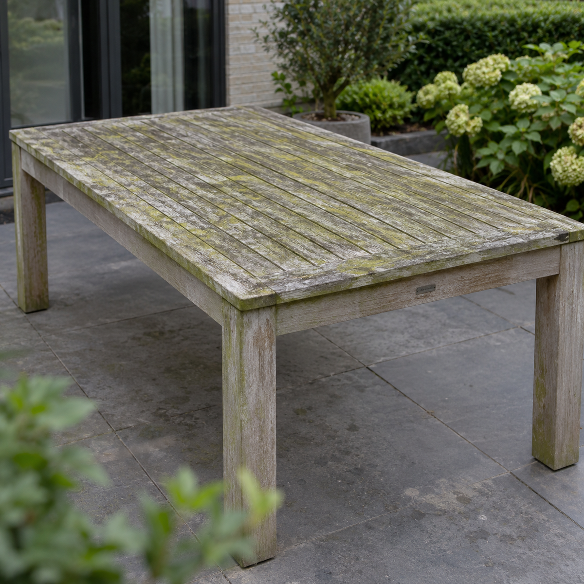 Illustratie van een verweerde teakhouten tuintafel met vergrijzing en groene aanslag, vóór reiniging.